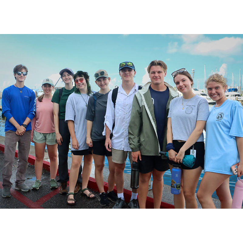 A group of 9 students are standing along a seawall next to the water.